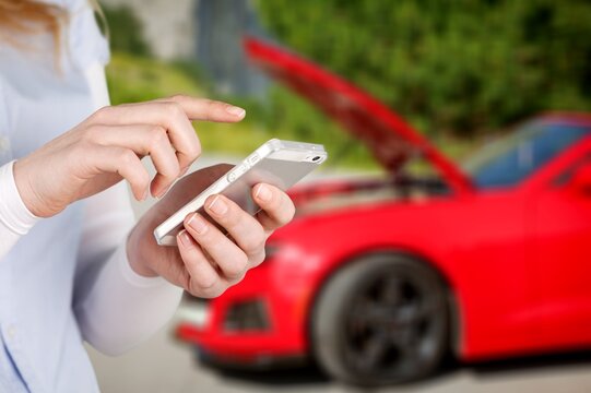 A woman using a smartphone to call for roadside assistance near a broken red car