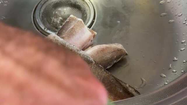 Pieces of hake fish defrosting and rinsing under running water in sink during homemade seafood preparation with natural kitchen atmosphere and fresh cooking process.
