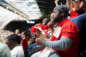 Naklejka premium Black fan does live betting during football game at the stadium filled with anticipation and excitement. Supporter check live odds on smartphone, tension and high risk of sports betting live.