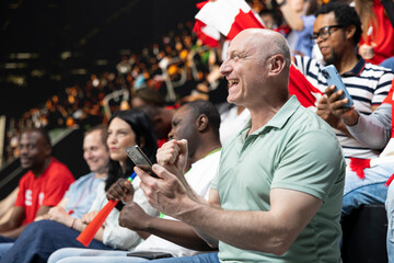 Obraz premium Stadium supporter checking online betting apps and live odds on phone, watching dramatic football match with diverse crowd of cheering fans. Enthusiast follow high risk live betting, bet ticket.