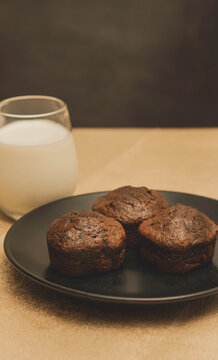 Stack of chocolate muffins on a black plate on a gold metal and black background with a glass of milk