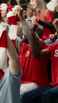 Vertical Video Excited football supporters celebrating with confetti in stadium stands, waving red scarves and cheering during live soccer match victory.