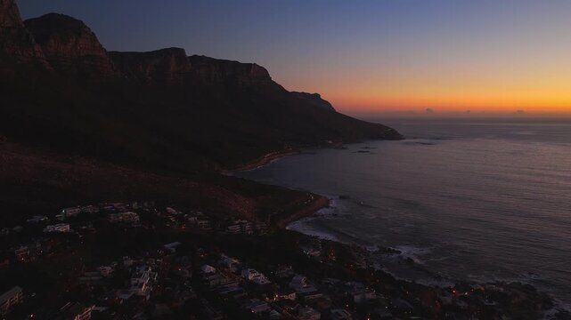 Aerial dusk scene in Cape Town shows Table Mountain and Twelve Apostles by Camps Bay as village lights glow, waves lap the curving coast, and the ocean surface darkens.