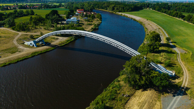 Aerial view of a modern arched gas pipeline bridge crossing the Vltava river surrounded by green fields and lush forests in Nova Ves, Central Bohemian Region, Czechia.