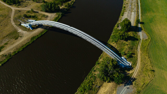 Aerial view of the white arched gas pipeline bridge crossing the Vltava river surrounded by green fields and dirt paths in Nova Ves, Central Bohemian Region, Czechia.