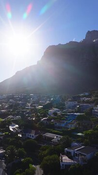 Aerial view of villas and pools beneath Table Mountain in Cape Town. The sun blazes above the ridge, lens flares streak, and the camera sweeps over winding roads.