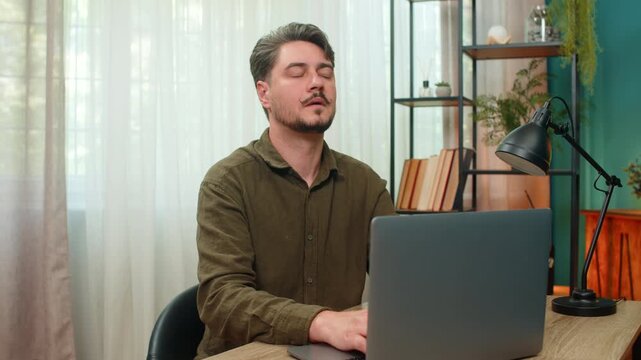 Middle-aged man at home finishes laptop tasks closes eyes sighs exhaustion seeking calm relief. Young adult guy at table exhales deeply feeling drained tired hoping rest soon revives strength again