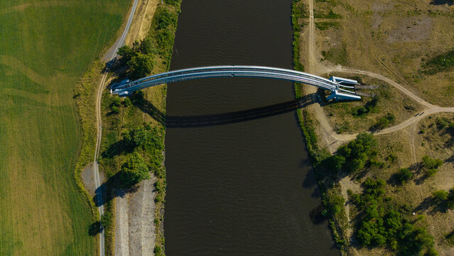 Aerial view of the Plynovod gas pipeline bridge crossing a river with its shadow on the water and surrounding green fields in Nova Ves, Central Bohemian Region, Czechia.
