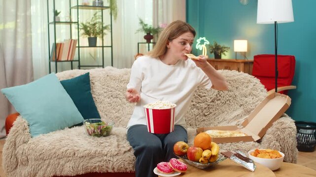 Pregnant middle-aged woman at home looks at pizza chocolate fruit chips popcorn eyes wide belly grumbles craving big feast. Caucasian girl on sofa plans small bites to calm hunger and keep baby safe.