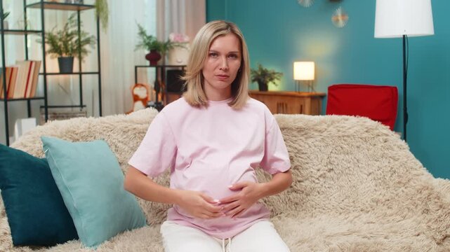 Pregnant middle-aged woman at home shows thumbs down after losing dislike gesture feeling frustration. Caucasian girl on sofa shakes head giving negative feedback sadness rejection seeking support, no