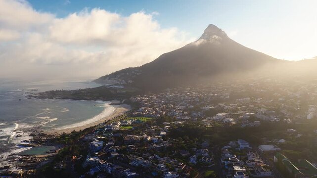 Aerial view of Cape Town at sunset shows Lion's Head, Camps Bay and Clifton, rocky shorelines, surf, rock pools, and palm promenades as a drone moves slowly.
