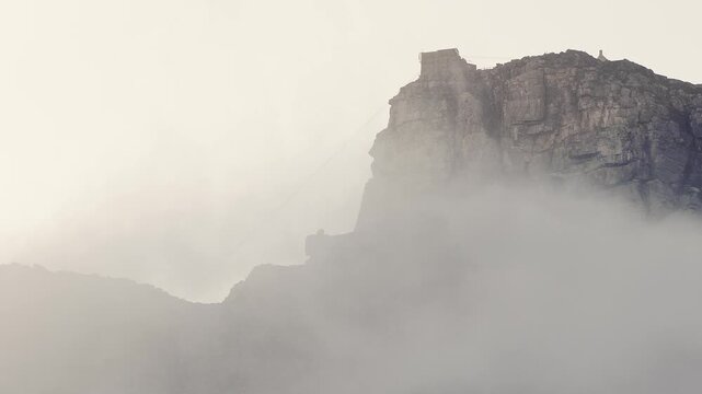 Table Mountain and cable car station in Cape Town sit in rolling mist. Soft backlight and wide framing reveal rugged cliffs and faint cables in slow fog.