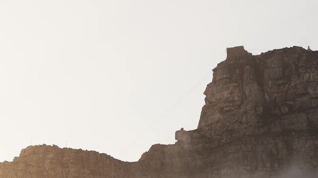 Rugged Table Mountain cliffs in Cape Town show the summit cableway station and diagonal cables, layered sandstone, warm low sun, soft mist, and a small peak building.