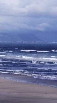 Aerial view of Cape Town coast shows empty sandy beach, rows of breaking waves toward Table Mountain across False Bay, storm clouds, wind driven surf, shorebirds.