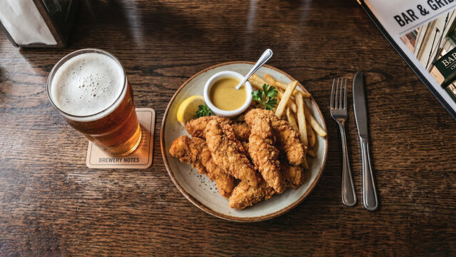Overhead view of a pub meal featuring crispy chicken tenders, french fries, mustard sauce, and a frothy pint of beer on a wooden table.