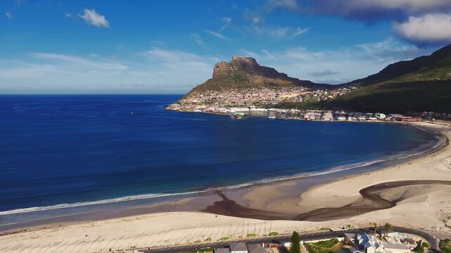 Aerial daytime view of Hout Bay, Cape Town, South Africa. Drone sweeps over deep ocean, pale sand, tidal stream, tiny figures, parked cars, and moored fishing boats.