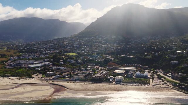 Aerial view of Camps Bay, Cape Town shows sandy beach, tidal channel, surf, beachgoers, cars, and the Twelve Apostles as sunbeams cast long late afternoon shadows.