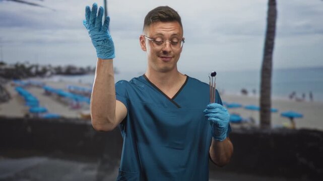 Man dentist in blue scrubs holding dental mirror instruments with gloved hand and hand to forehead gesture at a beach; amusement.