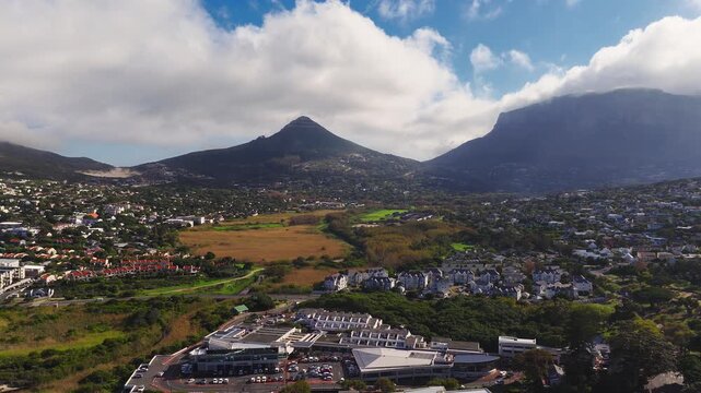 Aerial view of Cape Town, South Africa, with Lion's Head and Table Mountain as clouds spill over. Suburbs, a complex, and wetlands sit below in shifting light.