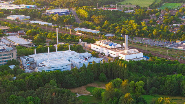 Aerial view of the GSK Stevenage pharmaceutical research and development site surrounded by lush green trees and a railway line during golden hour in Stevenage, England, United Kingdom.