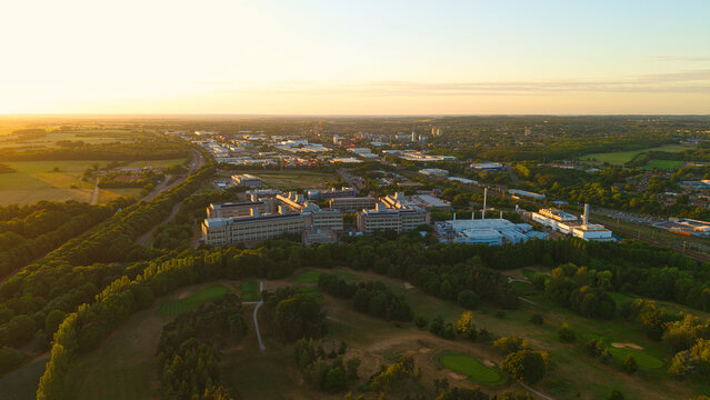 Aerial view of the GSK Stevenage research and development site surrounded by lush green trees and the urban landscape during golden hour in Stevenage, England, United Kingdom.