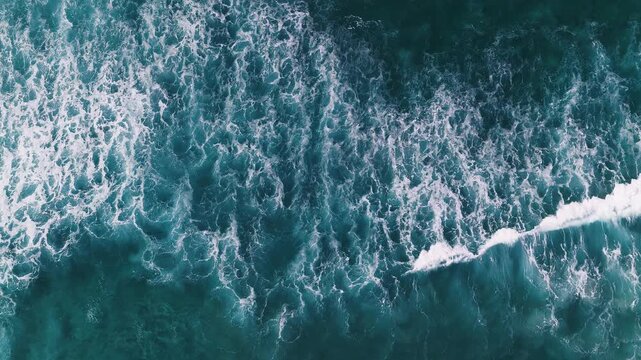 Aerial top down view of churning ocean near Cape Town. Multiple waves break and collapse into white foam, forming lace like patterns in daylight swell.