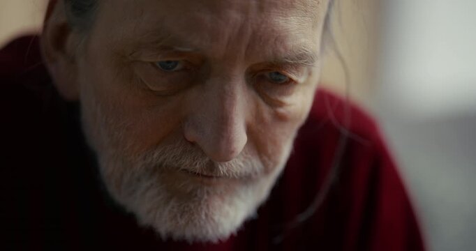 Pensive elderly man with gray beard looking down in soft indoor light