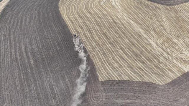 birdview of farmland where a tractor is driving to clean crops and prepare earth, create a dust clouds, and divides land diagonally into two distinguish sections.	