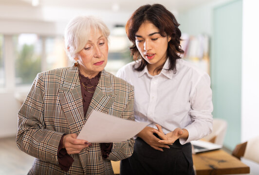 Senior woman and Asian girl colleagues review documents, read contract, discuss and resolve work issues, office hours. Mentoring and helping new young employees