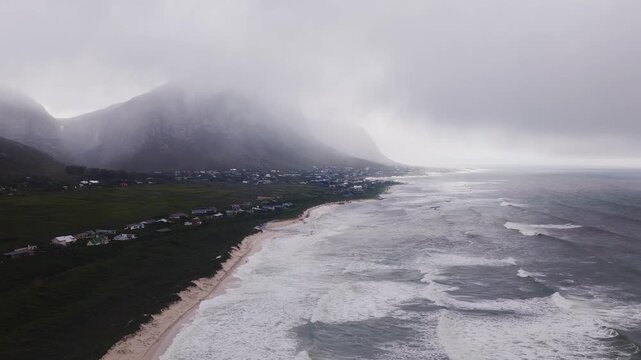 Aerial view of Cape Town Cape Peninsula shows curving sandy beach, coastal houses, rugged mountains under heavy overcast, waves rush ashore, fog drifts