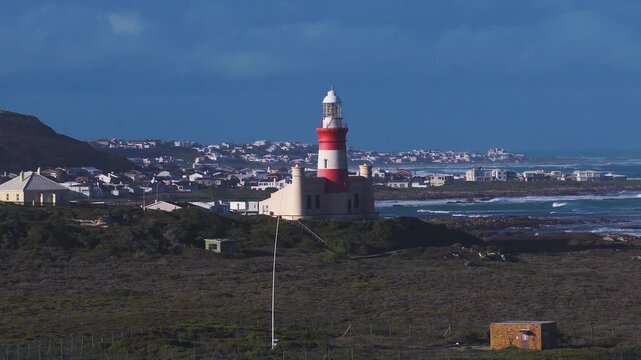 Aerial view of Cape Agulhas Lighthouse on a rocky promontory in South Africa. Surf rolls in, waves break, wind swept scrub and white houses sit under low side lighting.