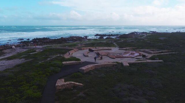 Aerial view of Cape of Good Hope star stone platform in Cape Town. Waves break, wind sweeps scrub, visitors walk paths. Wide panorama, cool tones, daylight shifts.