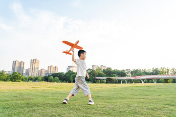 Obraz premium An East Asian young boy holds an orange toy airplane while walking on a spacious grassy park field under a bright daytime sky.