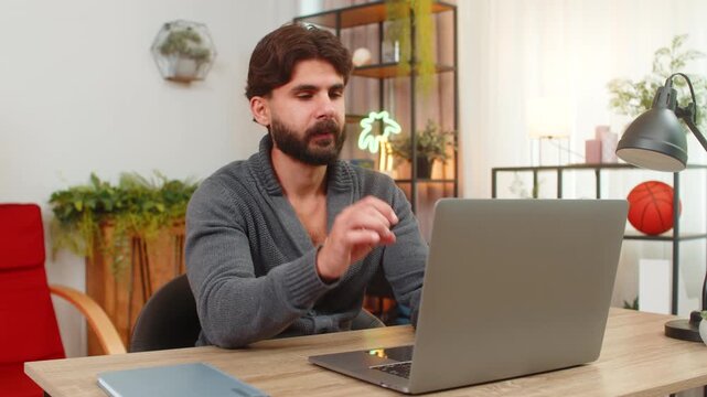 Indian man at home types details on laptop fills digital contract adds electronic signature breathes out relief. Arabian guy at desk shows approval hand sign smiles calm mood task done order confirmed