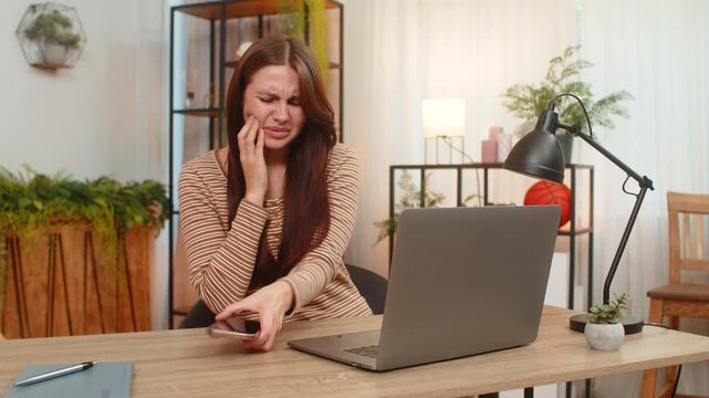 Young woman at home touches cheek feels tooth pain opens smartphone and searches dentist number. Caucasian girl at desk books visit online now types message worried about health and seeks swift relief