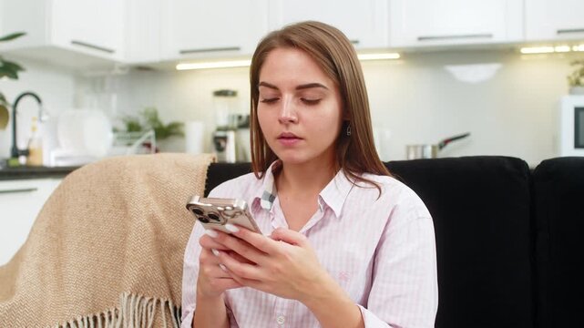 Young woman at home works on smartphone chat answers client types fast with thumbs focused on tasks and goals. Caucasian girl on sofa shows serious look strong focus customer support reply quick text.
