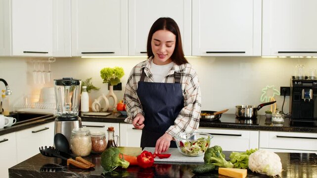 Young woman chops cherry tomatoes and prepares veggie salad on kitchen countertop for ketogenic diet lunch. Clean eating concept with fresh vegetables, apron, and home cooking routine, low carb lunch.