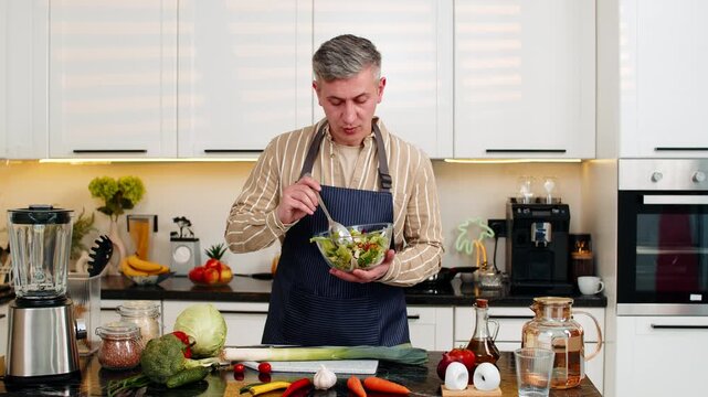 Middle-aged man holding vegetable salad in kitchen while giving diet advice and sharing nutrition ideas for home meals. Young adult guy looks cheerful and confident during healthy eating presentation.