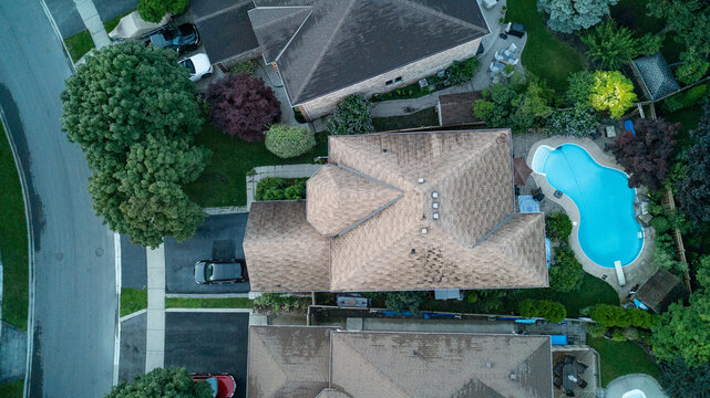 A direct top-down drone photograph of a detached residential home showing a weathered brown asphalt shingle roof, a paved driveway with a car, and a kidney-shaped swimming pool in the backyard.