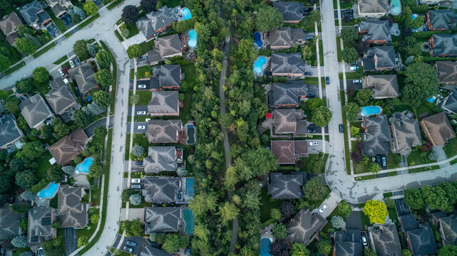 A vertical high-angle drone photograph looking down at two rows of suburban residential homes and backyard swimming pools divided down the center by a dense green belt of trees and a walking trail.