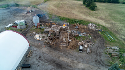 A wide aerial drone photograph of a collapsed wooden barn structure actively burning in a controlled demolition on a rural farm plot, surrounded by debris piles, silos, and heavy machinery. © Matt