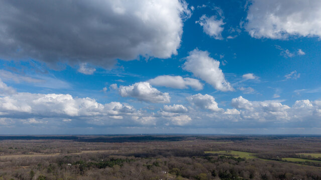 A high-resolution aerial view of rural North Mississippi featuring a mix of open farmland and dense woodland stretching to the horizon beneath a sky filled with large, well-defined cumulus clouds. 