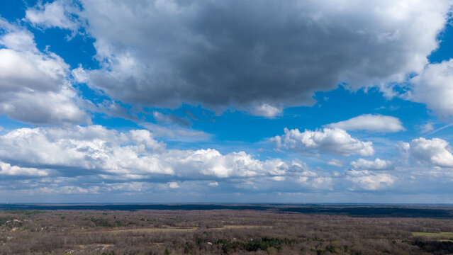 A high-resolution aerial view of rural North Mississippi featuring a mix of open farmland and dense woodland stretching to the horizon beneath a sky filled with large, well-defined cumulus clouds. 