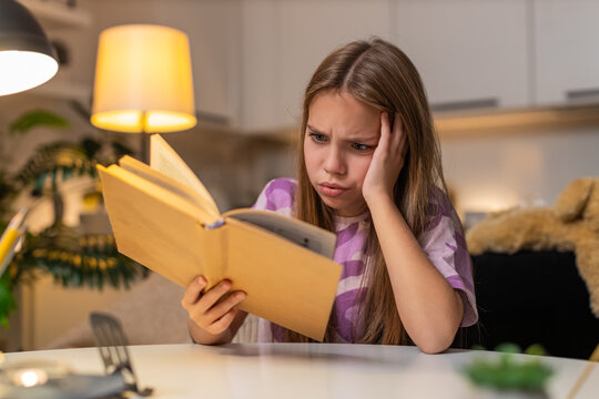 Teenager girl at night home reads page in book does not get task sighs feels upset holds head. Caucasian child kid sitting at table at evening looks at textbook frowns tries to solve problem slowly.