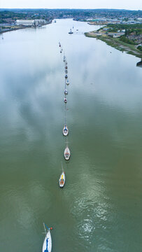Aerial view of sailboats moored in a straight line along the River Medway with industrial warehouses and green banks under a cloudy sky in Rochester, England, United Kingdom.