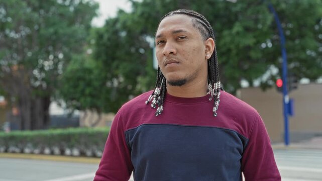 Man with beaded braids looks at camera while standing on a street crosswalk near trees and traffic light; quiet confidence.