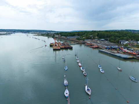 Aerial view of sailboats moored on the River Medway near industrial docks and lush green trees under an overcast sky in Rochester, England, United Kingdom.