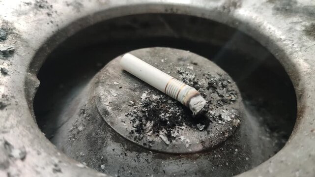 Smoldering Cigarette Butt in Dirty Metal Ashtray Close-Up