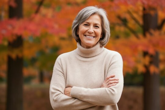 Mature woman with grey hair smiles warmly outdoors in park with colorful autumn foliage. Wears beige turtleneck sweater, arms crossed confidently. Image represents wisdom, contentment in later life.