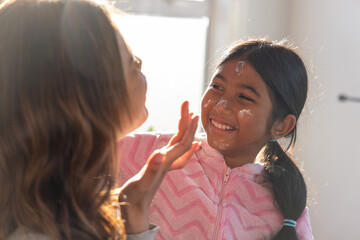 Mother gently smearing cream on Asian child wearing pink fleece at home near sunlit window © wavebreak3